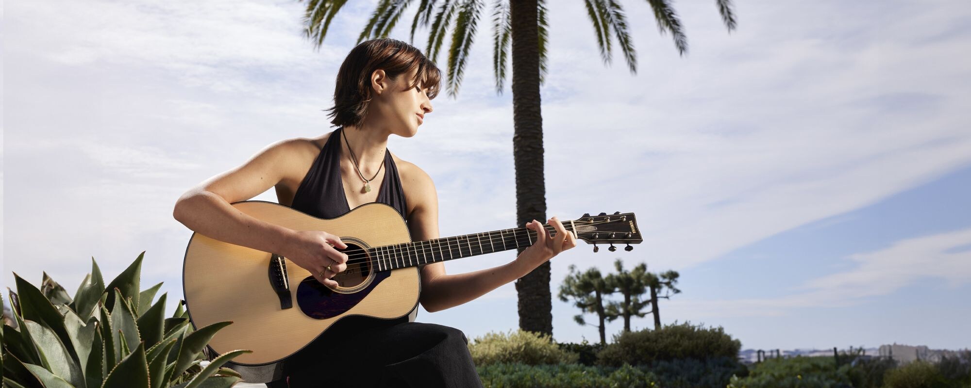 Guitarist Sophia James playing an FS9 guitar sits in front of a palm tree outside and plays an FS9 acoustic guitar.